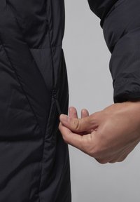 Sort puffer-jakke med struktureret overflade; nærbillede af en hånd, der justerer lynlåsen på siden; har et strømlinet design og ergonomisk pasform.