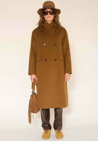 Manteau croisé en laine marron avec des boutons noirs, associé à un pantalon en cuir marron et des baskets en suède jaune. Accessoirisé avec un chapeau et un sac marron.