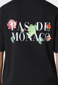 Černé tričko s bílým textem "PAS DE MONACO," obklopené barevnými květinovými vzory. Krátké rukávy a volný střih.