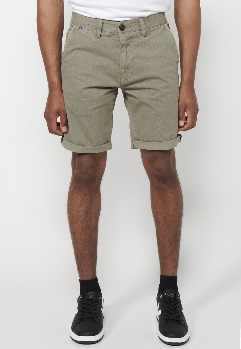 Man in opgerolde olijfgroene shorts, zwarte sokken en zwarte sneakers die tegen een witte achtergrond staat.
