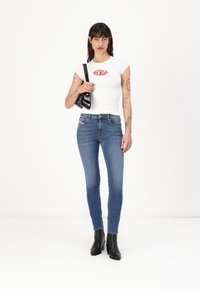 Camiseta blanca ajustada con logo rojo, jeans ajustados azules y botas negras hasta el tobillo. Modelo sosteniendo un pequeño bolso negro.