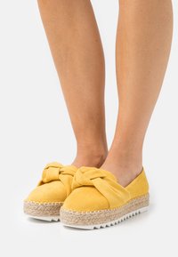 Le espadrillas in suede giallo presentano un design con upper annodato, una suola intermedia intrecciata in juta e una suola esterna in gomma bianca con un bordo seghettato.