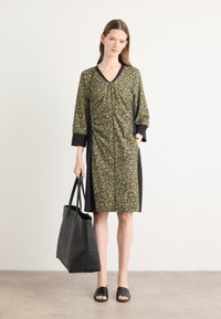 Oliivinvihreä mekko leopardikuviolla, mustat hihat ja sivupaneelit. Malli pitää mustaa olkalaukkua ja käyttää mustia liukusandaaleja.