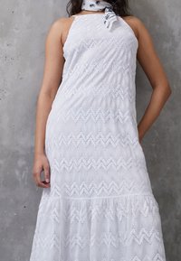 Robe sans manches en dentelle blanche avec des motifs géométriques, portée avec un foulard de cou blanc et noir à motif paisley devant un mur en béton gris.