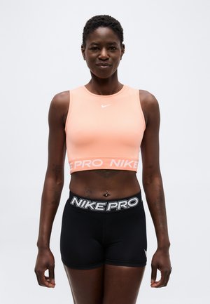 Personne athlétique portant un crop top pêche et un short noir Nike Pro, debout devant un fond clair uni.
