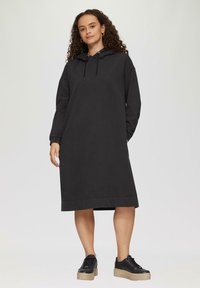 Czarna sukienka z kapturem i długim rękawem, o oversize'owym kroju, z prążkowanymi mankietami i prostym dołem, w zestawie z czarnymi trampkami na platformie.
