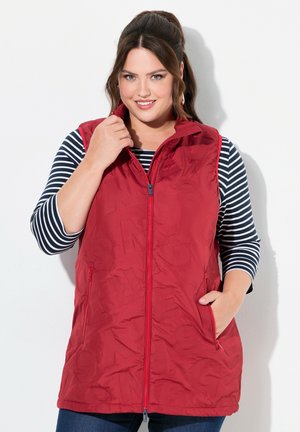Chaleco acolchado rojo con cuello alto, cremallera frontal y bolsillos laterales, llevado sobre una camiseta de manga larga a rayas en azul y blanco.