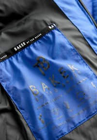 Unutrašnjost crne jakne s plavom oznakom koja ima ugravirani logo "BAKER by TED BAKER", simbole za njegu i tekstualne informacije o materijalima i značajkama.