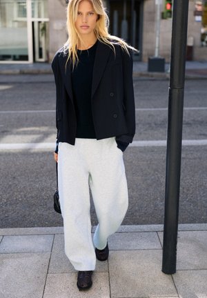 Une femme blonde se tient sur un trottoir en ville. Elle porte un blazer noir, un haut foncé, un pantalon blanc et des mocassins, et tient un petit sac à main noir.