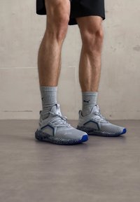 Gray athletic shoes with blue accents, textured soles, and a mesh upper, worn with gray striped socks and black shorts against a light background.