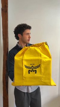 Bărbat cu păr creț și închis la culoare ține o geantă mare galbenă cu șnur MCM în fața unui perete alb simplu.