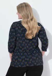 Frau mit langen blonden Haaren, die ein schwarzes Oberteil mit blauen und beigen vertikalen Streifen sowie schwarze Hosen trägt, vor einer weißen Wand steht und die Hände in den Taschen hat.