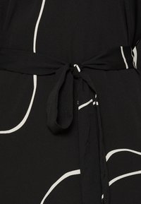 Ceinture en tissu noir nouée en un nœud sur un vêtement noir orné de lignes courbes abstraites blanches.