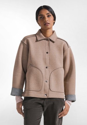 Donna che indossa una giacca beige con bottoni, grandi tasche frontali e polsini grigi risvoltati, abbinata a pantaloni scuri in velluto a coste.