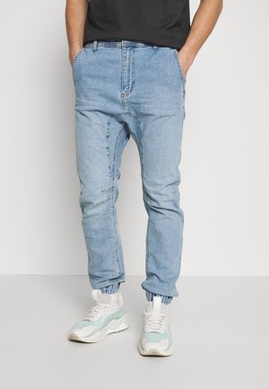 Mann trägt hellblaue Denim-Jogger-Jeans mit elastischen Bündchen und weiß-grünen Turnschuhen, Hände in den Taschen, steht vor einem einfarbigen Hintergrund.