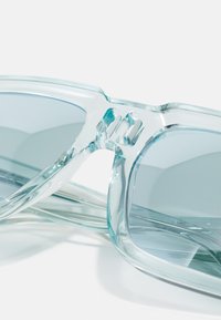 Ochelari de soare supradimensionați, transparenti, cu un cadru albastru translucent și lentile gri. Designul unic include o legătură la punte și colțuri rotunjite.