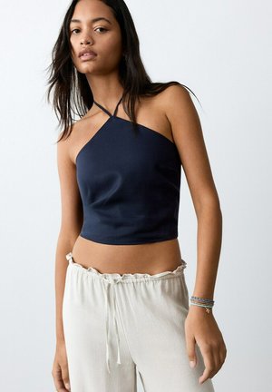 Joven con un top corto azul marino de tipo halter y pantalones beige claros con cordón, con pulseras en la muñeca izquierda, sobre un fondo liso.