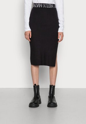 Persoon draagt een zwarte Calvin Klein geribbelde midi-rok met logoband, wit topje met lange mouwen en zwarte enkellaarzen, staand op een grijze vloer.