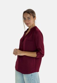 Blusa bordeaux con un design a bottoni, dettagli con tasca, maniche arrotolabili e tessuto leggero, abbinata a jeans azzurri.