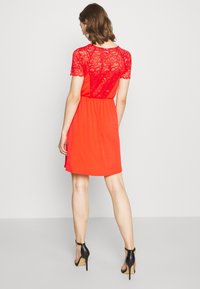 Czerwona sukienka z koronkową górą, krótkimi rękawami i zwiewną spódnicą. Tył wykonany jest z gładkiej tkaniny z taliowaniem, a dół sięga ponad kolana.