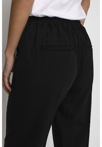 Pantalons noirs à taille élastique avec une texture lisse, dotés de deux poches arrière et d'une coupe décontractée. Le tissu semble léger et respirant.