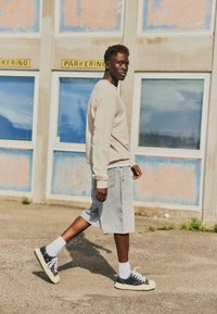 Jeune homme marchant en plein air près d'un bâtiment usé, portant un sweat-shirt beige, un short gris, des chaussettes blanches et des baskets noires.