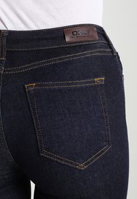 Mörkblå denimjeans med en slät yta, med gula sömmar och en lädermärkesetikett på midjan. En bakficka synlig.