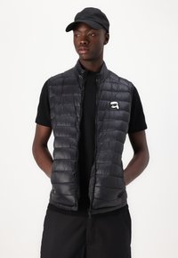 Melnā pūkainā veste ar quilted tekstūru, ar priekšējo rāvējslēdzēju un sānu kabatām, uzvilkta virs melnas īsām piedurknēm krekla, kuram ir balta logo.