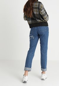 Zwart-geel geruite cropped trui, blauwe distressed denim jeans met rafelige manchetten en witte sportschoenen.