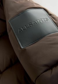 Brūns pildīts jakas ar teksturētu melnu ādas ieliktni, uz kura ir iepresēts "ALLSAINTS" logo. Gluda auduma ar izšuvumu detaļām.