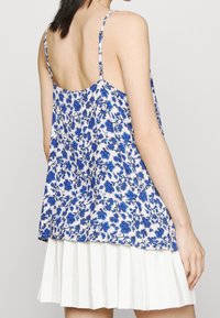 Kvinna i en ärmlös, lös vit topp med blå blommönster och en plisserad vit kjol, sedd från ryggen mot en enkel bakgrund.