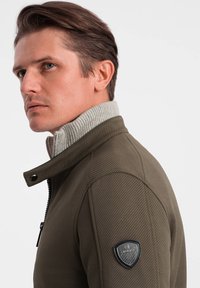 Giacca verde oliva con collare a costine grigio, tessuto testurizzato, chiusura con zip e una patch nera con logo sulla spalla. Design slim fit.