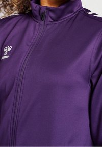 Lila zip-up jacka gjord av smidigt, stretchigt tyg; har hög krage och svarta accenter på axlarna; logotyp på vänster sida.