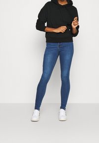 Sudadera con capucha negra y jeans ajustados azules, combinados con zapatillas blancas. La sudadera tiene un bolsillo en el frente y cordones.