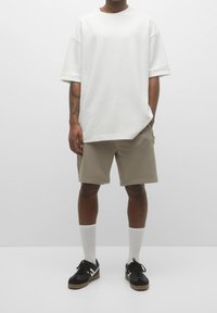 T-shirt blanc surdimensionné à manches courtes, short beige, chaussettes blanches montantes jusqu'aux genoux, baskets noires avec accents beiges. Matières texturées unies.