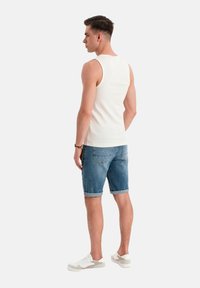 Witte tanktop, spijkerbroek shorts met opgerolde mouwen en witte sneakers met grijze accenten. Model wordt van achteren getoond.