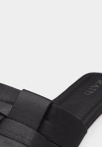 Μαύρο δερμάτινο σανδάλι τύπου slide με φαρδιές πλεκτές λουρίδες και επίπεδη σόλα σε κοντινή λήψη.