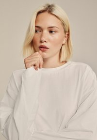 Witte lange mouwen t-shirt met een ronde halslijn, met een zachte stoftextuur. Het model heeft lichtblond haar en steunt een hand tegen haar gezicht.