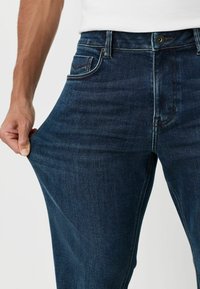 Dunkelblaue Denim-Jeans mit entspannter Passform, ausgestattet mit fünf Taschen, einem Reißverschluss und sichtbaren Steppnähten. Das Material hat einen leichten Stretch.