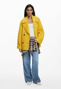 Gul dubbelknäppt ullkappa med spetsiga slag, kombinerad med en vit croptop och lösa ljusblå jeans. Rutig flanellskjorta knuten i midjan.