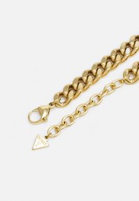 Gouden metalen kettingarmband met grote schakels en een karabijnsluiting. Kenmerkende driehoekige bedel met 'GUESS'-graveerwerk. Glad oppervlak.