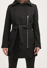 Mustan vyön kanssa varustettu villakangastakki, jossa on epäsymmetrinen etuvetoketju, päällä mustat housut, näkyy reiden puolivälistä olkapäihin.