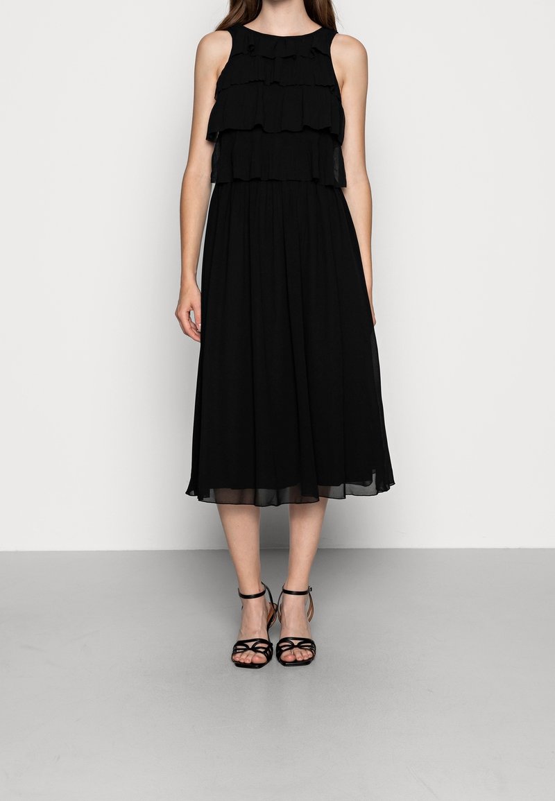 Femme portant une robe midi noire sans manches avec des volants superposés et des sandales noires à talons hauts avec des lanières, debout devant un fond blanc uni.