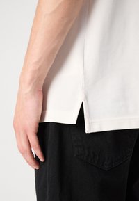 Polo à manches courtes beige clair en tissu texturé, avec des fentes latérales. Porté avec un pantalon noir avec une poche visible.
