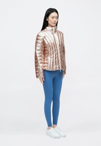 Fémes rózsaarany színű, függőleges steppelt mintájú, magas nyakú, cipzáras steppelt dzseki, amelyet kék leggingssel és fehér tornacipővel viselnek.