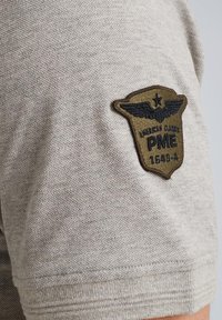 Camisa de manga corta beige con tejido texturizado y un parche bordado marrón oscuro que presenta alas, "PME" y acentos "1649-4".