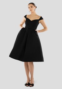 Zwarte strapless jurk met een aansluitend lijfje, gerende rok en zijzakken. De stof heeft een gladde textuur. Geaccentueerd met open teen schoenen.