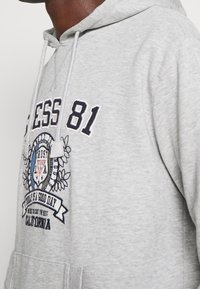Sudadera gris con un gráfico en azul marino que dice "LESS 81" y elementos decorativos. Incluye una capucha con cordones y un bolsillo canguro.