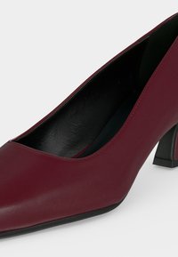 Escarpin en cuir bordeaux à talon moyen, bout rond et doublure intérieure noire sur fond blanc.