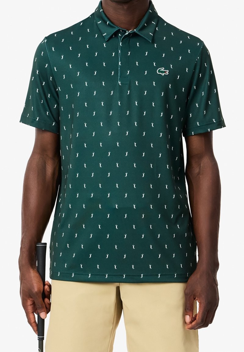 Polo vert avec de petites impressions blanches de golfeurs, manches courtes, col classique, et un logo Lacoste sur la poitrine gauche. Texture de tissu lisse.